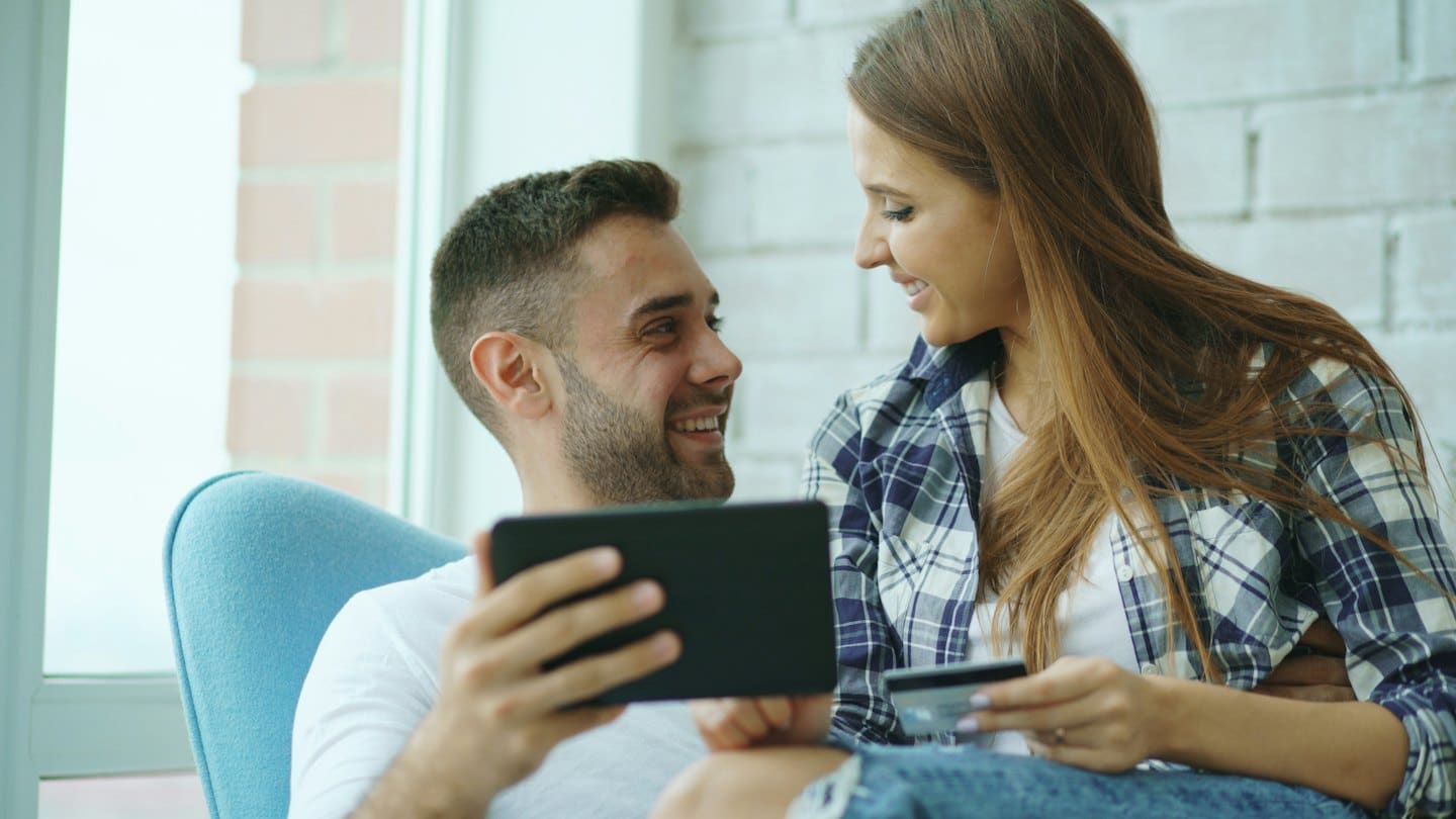 Coppia sorride mentre fa acquisti online con un tablet e una carta di credito.